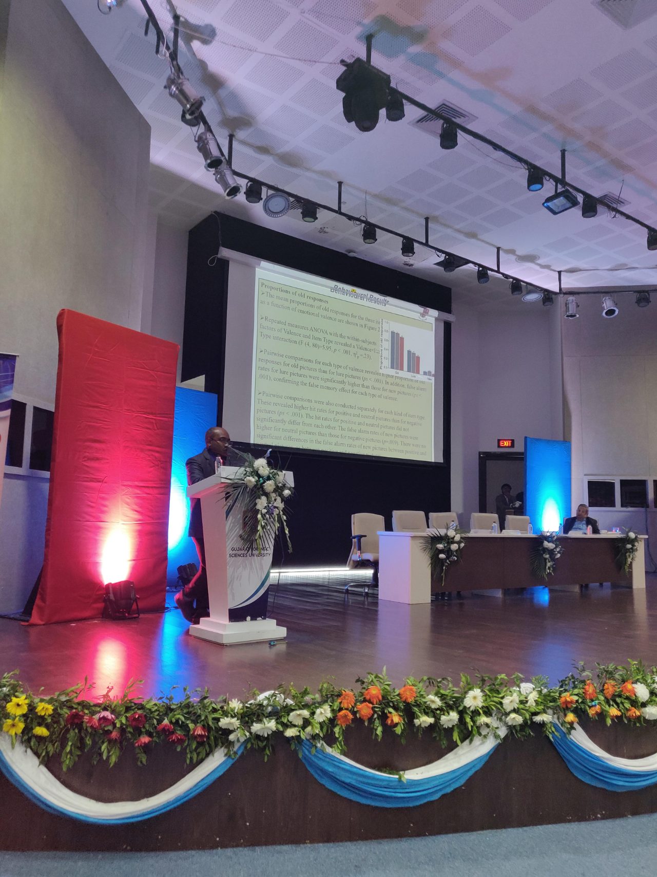 A speaker at the lectern beneath a slide of behavioural results on emotional valence and false memory