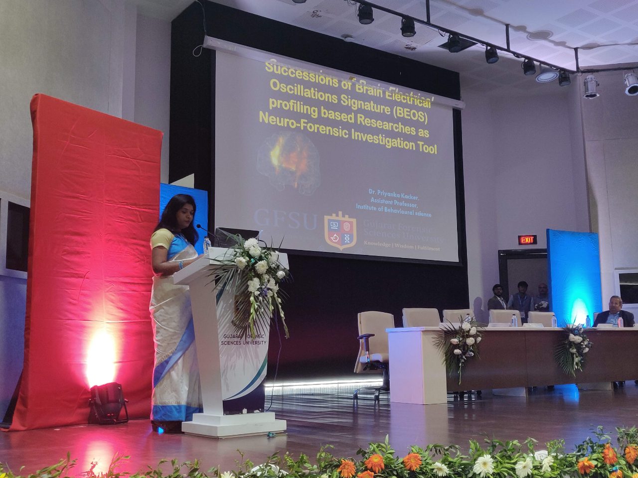 Dr Priyanka Kacker at the lectern, her title slide on BEOS profiling as a neuro-forensic investigation tool on the screen behind