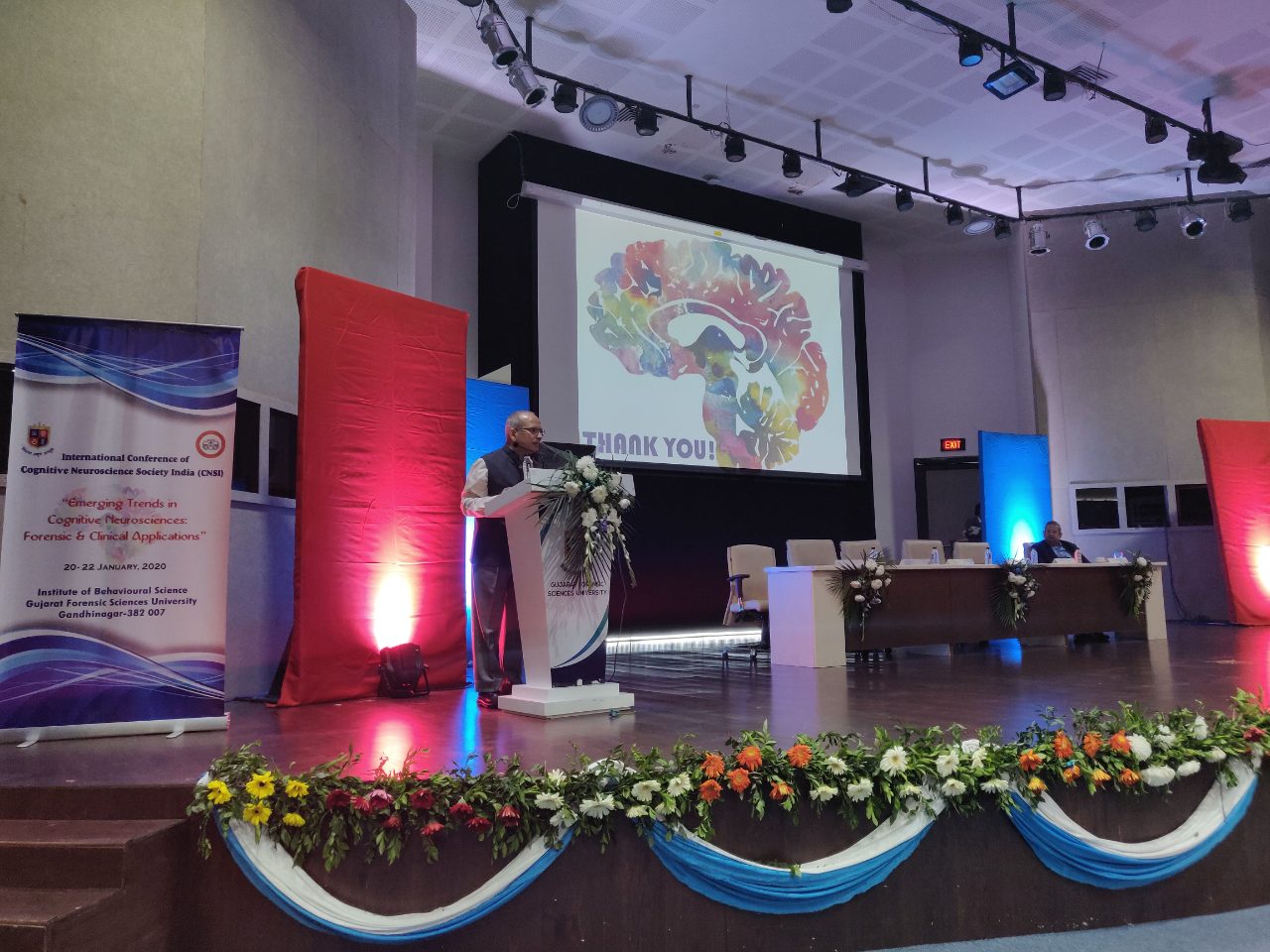 A speaker at the lectern of the GFSU Auditorium, the CNSI-2020 banner at left and a closing ‘Thank you!’ slide on the screen