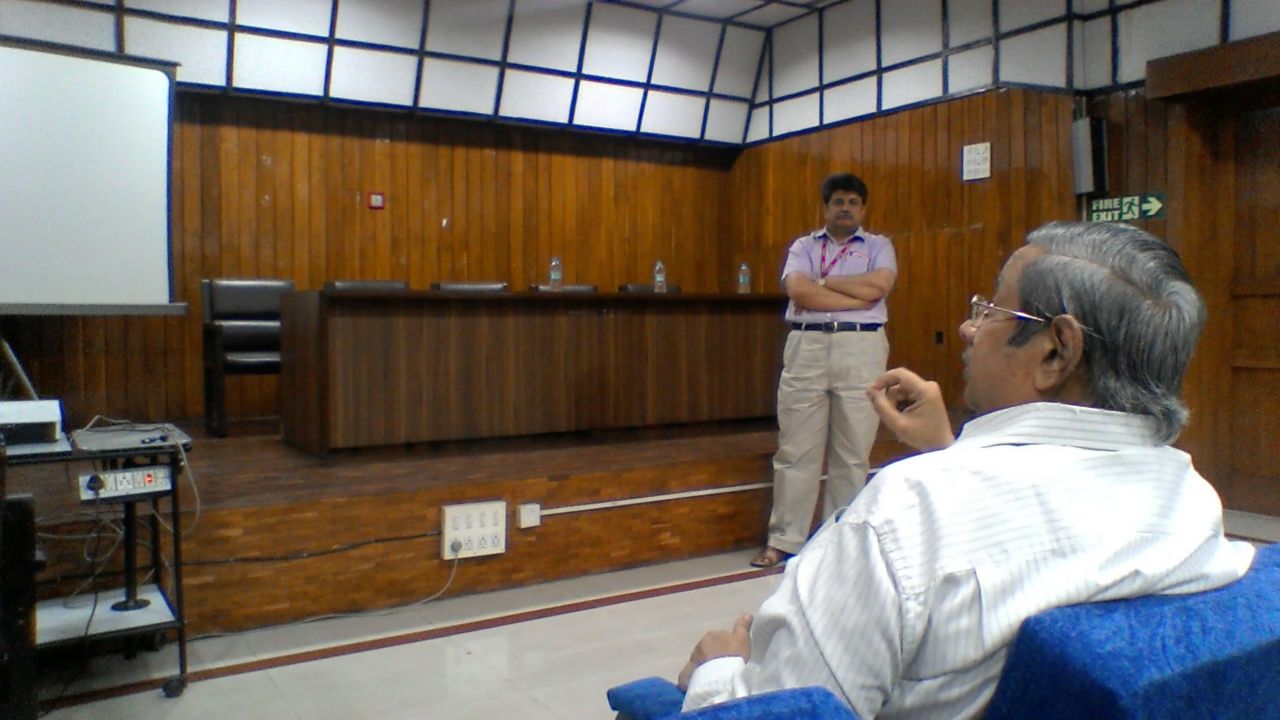 A man in a lilac shirt stands before the dais, arms folded, as a listener in the front row speaks