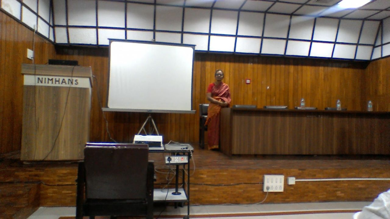 A speaker in a red sari stands by the dais, the NIMHANS lectern and the projection screen to the left