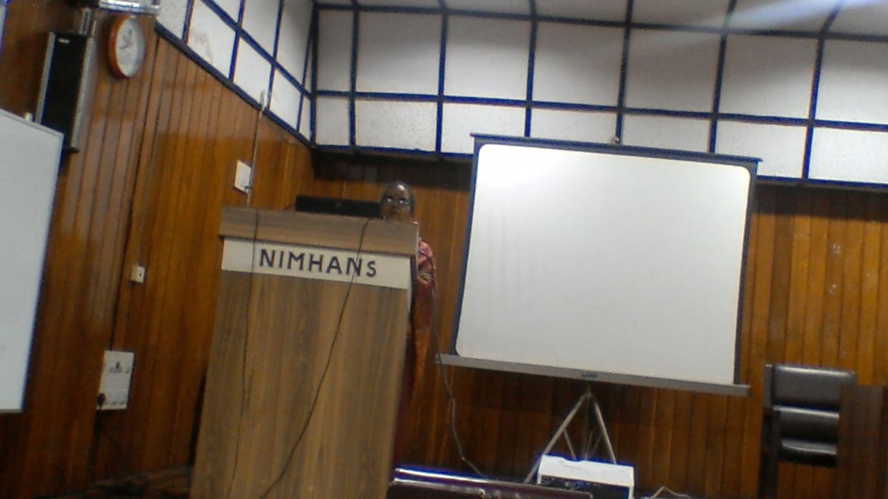 The same speaker at the NIMHANS lectern, the projection screen beside her