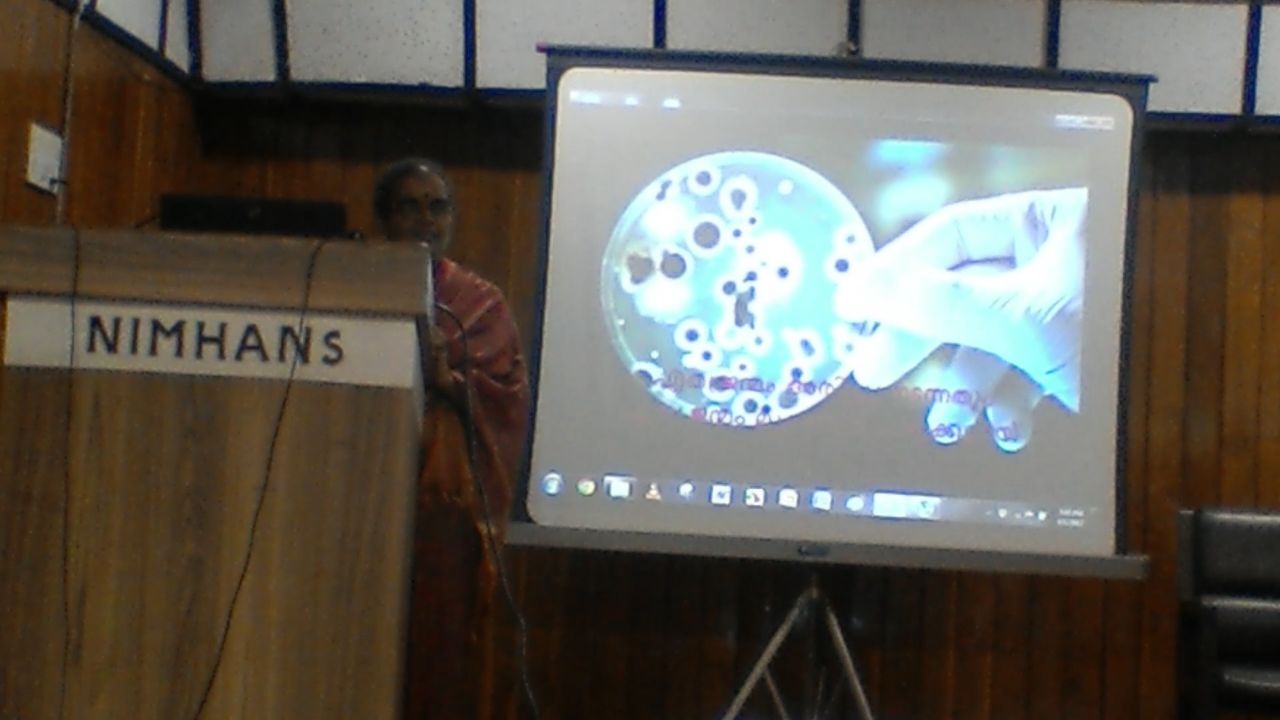 The same speaker at the lectern beside a projected image of a gloved hand holding a culture dish