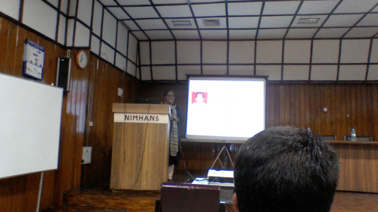 Another speaker smiles from the NIMHANS lectern beside a slide with a portrait photograph