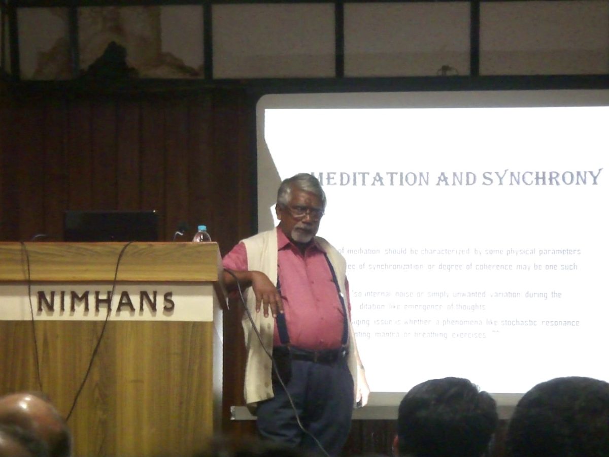 Prof. Sisir Roy beside the NIMHANS lectern, a slide on meditation and synchrony behind him, at the first Cognitive Neuroscience Forum, 28 January 2017